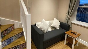 Un salon avec un canapé gris foncé, des coussins blancs, une petite table d’appoint en bois et une fenêtre avec des rideaux.