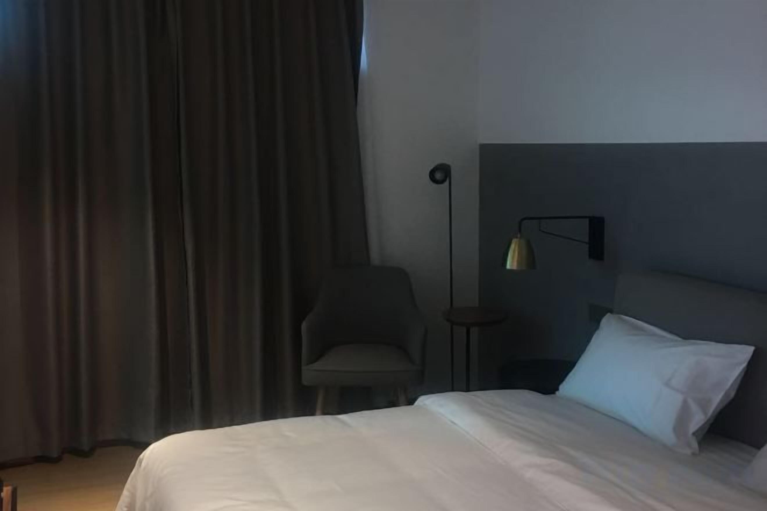 A hotel room with a bed, a chair, a bedside table, and a lamp.