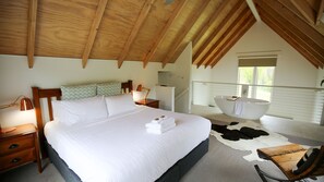 A bedroom with a large bed, a wooden ceiling, a window, a bathtub, and a cowhide rug. - Abby's Cottage - Bright (Wandiligong)