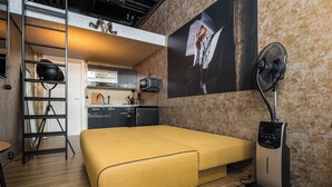 Habitación compacta con sofá cama, cinta de correr, ventilador de pared, zona de cocina y una escalera.