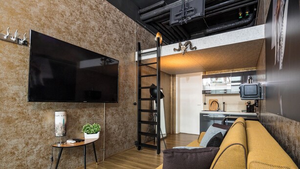 Una sala moderna con un televisor de pantalla plana, una mesita con una planta, un sofá amarillo y una zona de cocina con microondas y televisor.