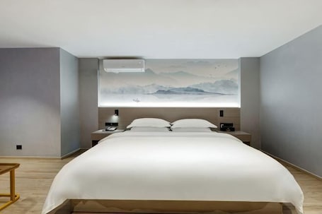 A modern hotel room with a large bed, a nightstand, a wall-mounted air conditioner, and a painting of mountains.. limu Hotel