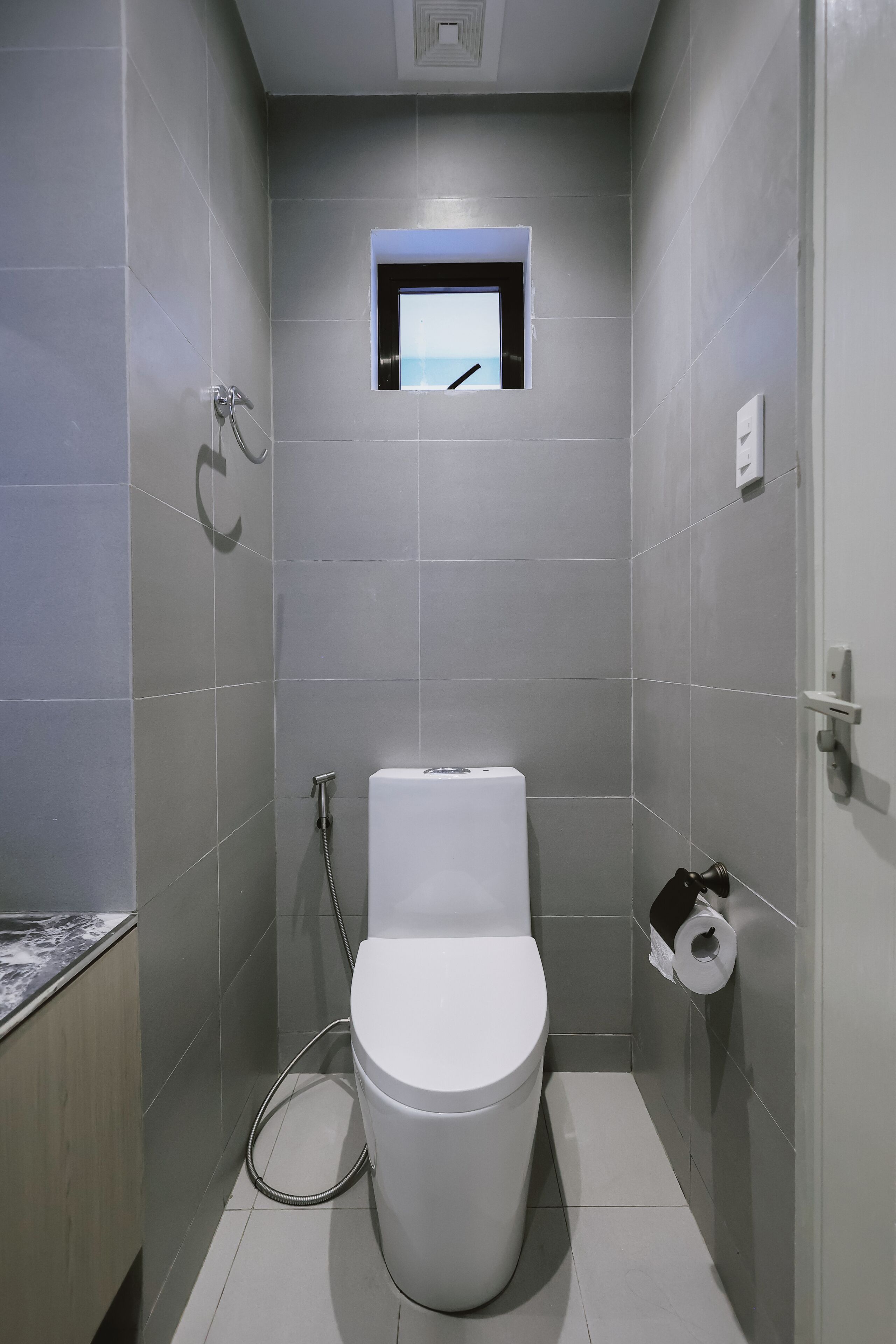 A small, modern bathroom with grey tiles, a toilet, a shower, and a small window.