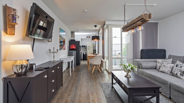 Ein stilvoller Wohnraum mit dunklem Holzboden, einem grauen Sofa mit Kissen im Zeitungsdesign, einem dunklen Holztisch und einem an der Wand montierten Fernseher.