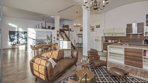 Un vaste espace de vie comprenant un canapé en cuir marron, une table basse en bois et un lustre. Un escalier mène à un niveau supérieur, et il y a également un coin cuisine avec des armoires blanches et un plan de travail en bois.