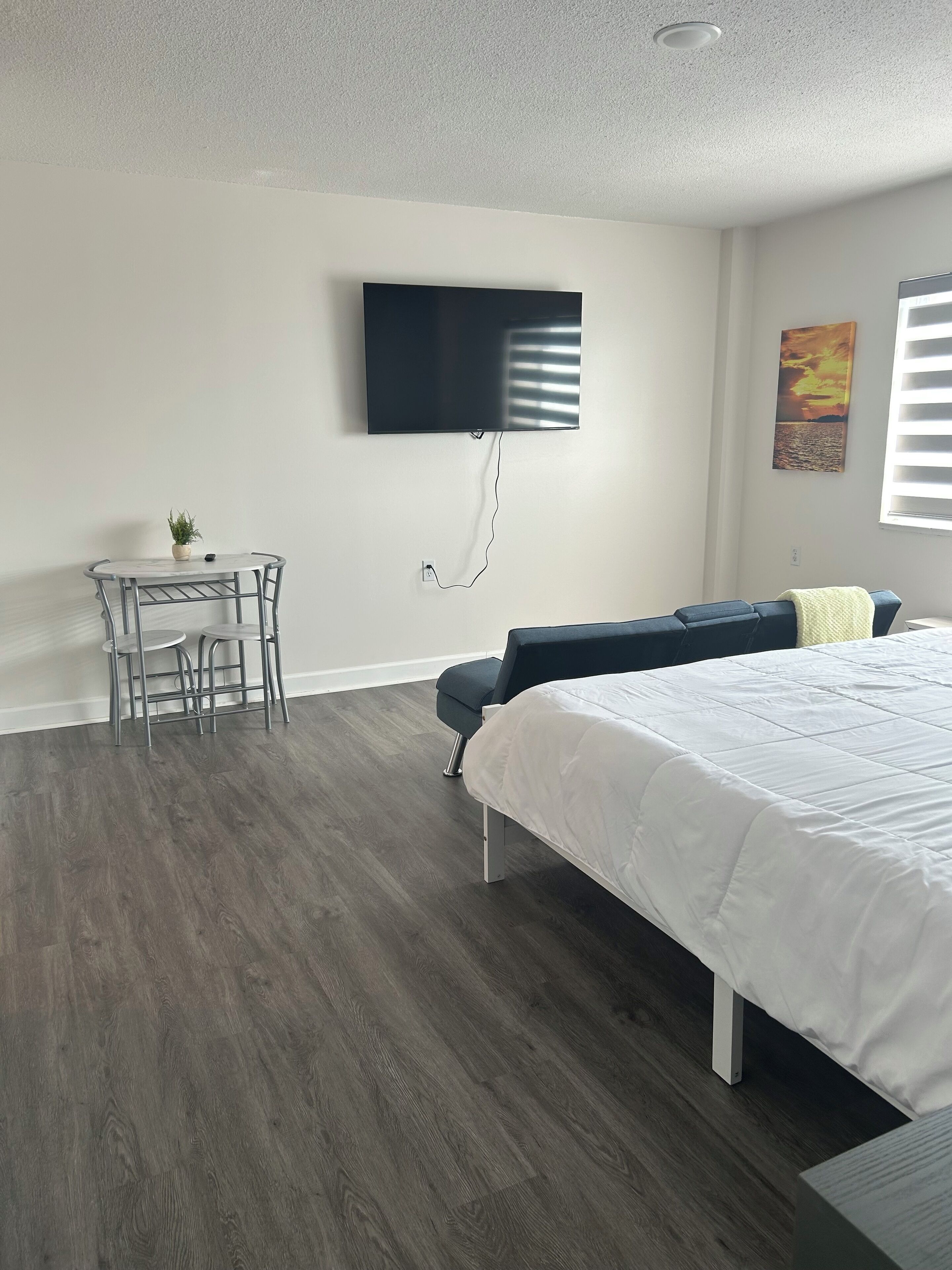 A modern bedroom with a bed, a TV mounted on the wall, a small table with chairs, and a window with blinds.
