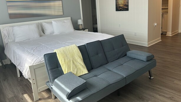 A bedroom with a bed, a gray sofa, a painting of a sunset, and a view into the kitchen. - Trendy apartment in Gadsden, AL (Gadsden)