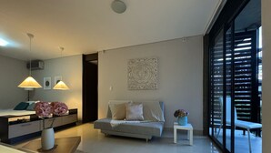 A modern living room with a sofa, a coffee table, and a side table with flowers. There is a large window with blinds, a ceiling light, and a wall-mounted artwork.