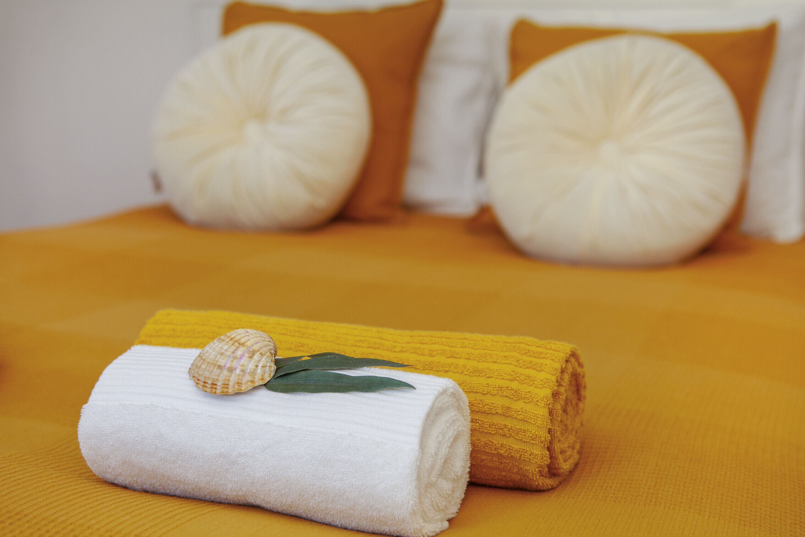 A neatly made bed with a yellow bedspread, white pillows, and a rolled white towel with a seashell and leaf.