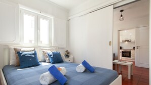 Un lit bien fait, avec une literie bleue et blanche, une table de chevet avec une lampe, et une vue sur la cuisine par une porte ouverte.