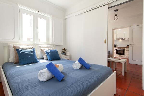 A neatly made bed with blue and white bedding, a bedside table with a lamp, and a view of the kitchen through an open door.