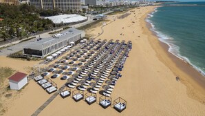 An aerial view of a beach with rows of sunbeds and umbrellas, a hotel with a large roof, and a coastline with buildings in the background.