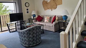 Een woonkamer met een bank, voetenbankje, salontafel en televisie. Er hangt een klok aan de muur en er is een raam met gordijnen.