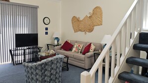 Een woonkamer met een bank, poef en een televisie. Er is een trap met een leuning, een wandklok en een wanddecoratie van geweven materiaal.