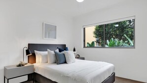 Une chambre à coucher moderne avec un grand lit, du linge de lit blanc, une table de chevet avec une lampe, un tableau encadré au mur et une fenêtre donnant sur un paysage verdoyant.