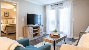 Una sala de estar con un televisor, una mesa de centro y dos sofás. Hay una puerta que da a un dormitorio con cama y ventilador de techo.