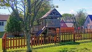 A wooden playhouse with a slide, surrounded by a wooden fence, set in a grassy yard with houses in the background. - Valley Lodge - UK49228 (Gunnislake)