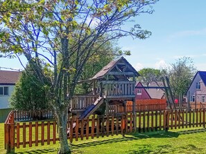 A wooden playhouse with a slide, surrounded by a wooden fence, set in a grassy yard with houses in the background. - Valley Lodge - UK49228 (Gunnislake)