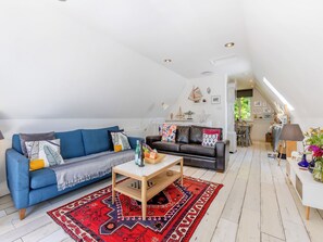A spacious loft-style living area with a blue sofa, a dark leather couch, and a wooden coffee table. A red patterned rug is on the floor, and there is a small table with a lamp and decorative items.