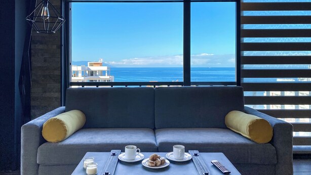 A modern room with a grey sofa, a small table with a plate of pastries, two cups, and a remote control. A large window offers a view of the sea and buildings.