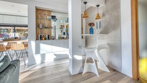 A modern kitchen with white cabinetry, a glass shelving unit, and a dining area with wicker chairs. - La Canopèa: The Art of Living in Cap Ferret, Between the Pine Forest and the Ocean (Lège-Cap-Ferret)