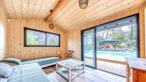 A wooden cabin interior with a bed, a sofa, a table, and a sliding glass door leading to an outdoor pool area. - La Canopèa: The Art of Living in Cap Ferret, Between the Pine Forest and the Ocean (Lège-Cap-Ferret)