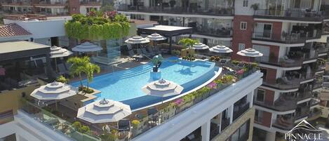 A rooftop pool with sun loungers and umbrellas, surrounded by balconies and greenery.