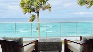 A balcony with two wooden chairs and a woven ottoman, overlooking a turquoise ocean with waves. - Blue Green (Sosúa)