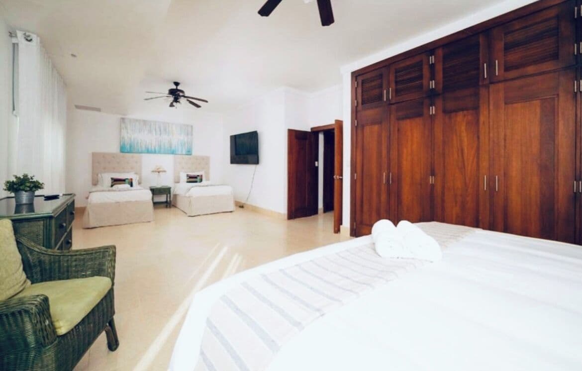 A bedroom with a large bed, a wooden wardrobe, a ceiling fan, and a seating area with a chair and a small table.