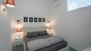 A bedroom with a bed, two bedside lamps, a wall-mounted air conditioner, and a window.