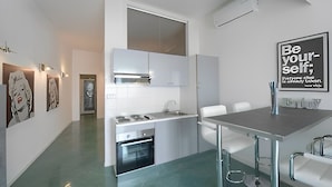 A modern kitchen with white cabinetry, a built-in oven, and a stainless steel countertop. There is a dining table with chairs, and a wall-mounted air conditioning unit.