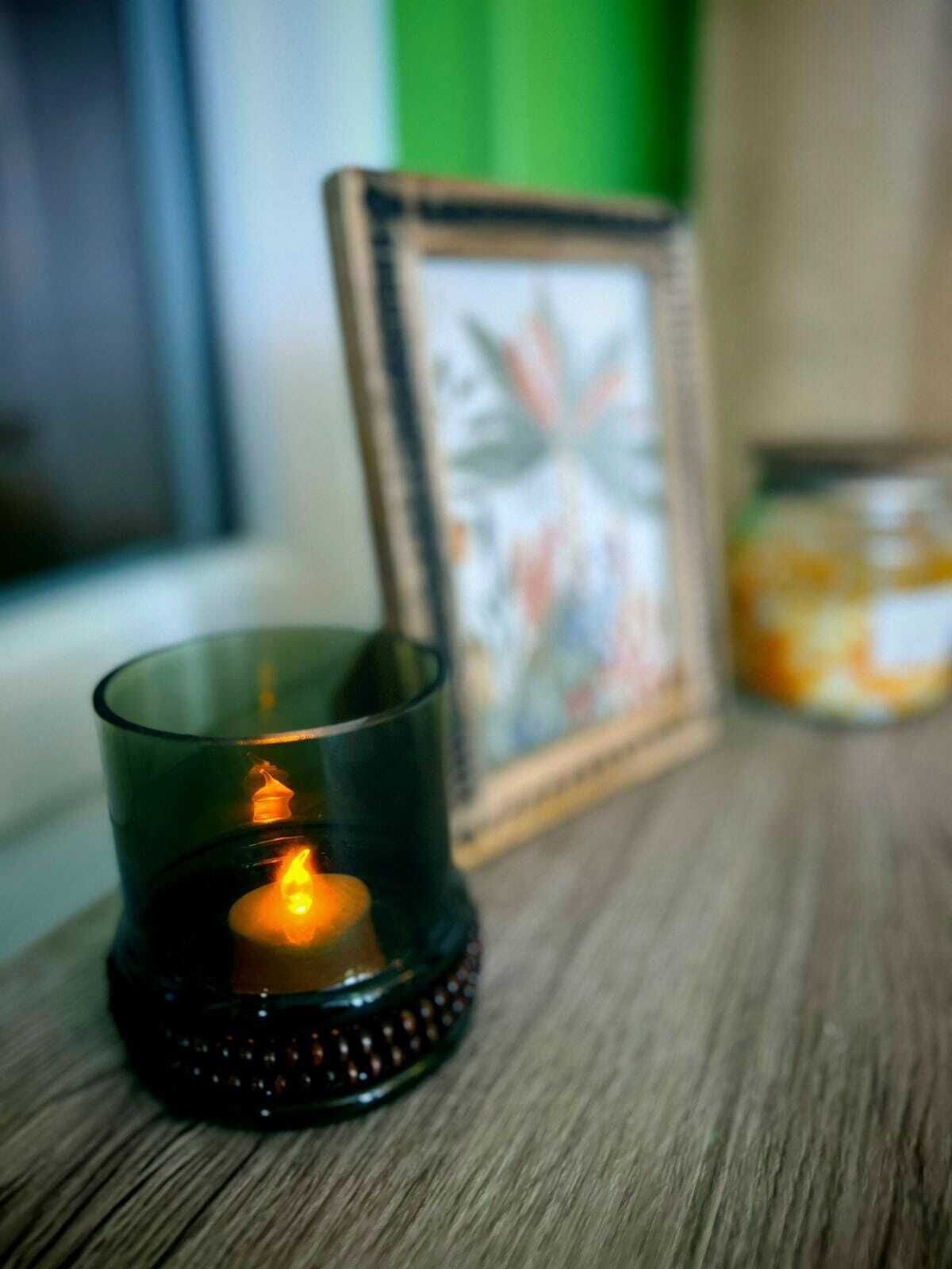 Une bougie dans un photophore en verre posé sur une surface en bois, un tableau encadré et un pot de bougies.