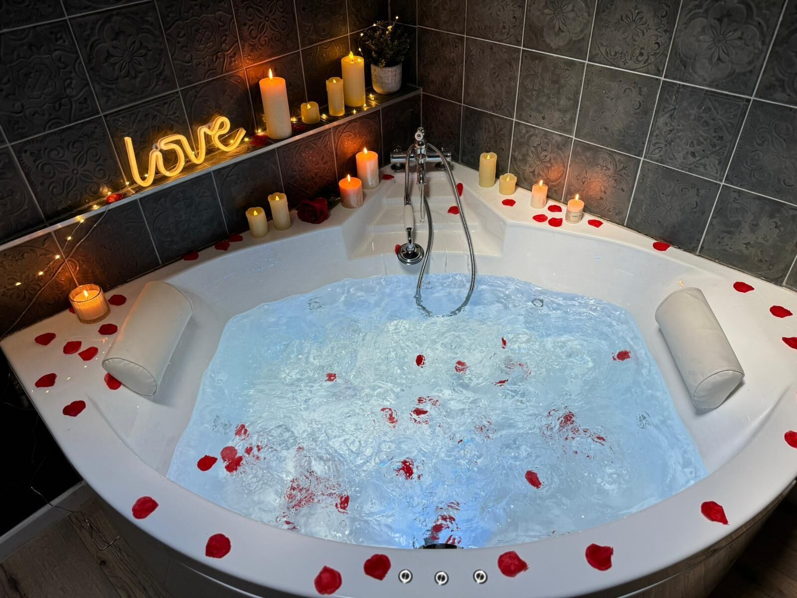 A bathtub filled with water and rose petals, surrounded by candles and a neon sign that reads "love".
