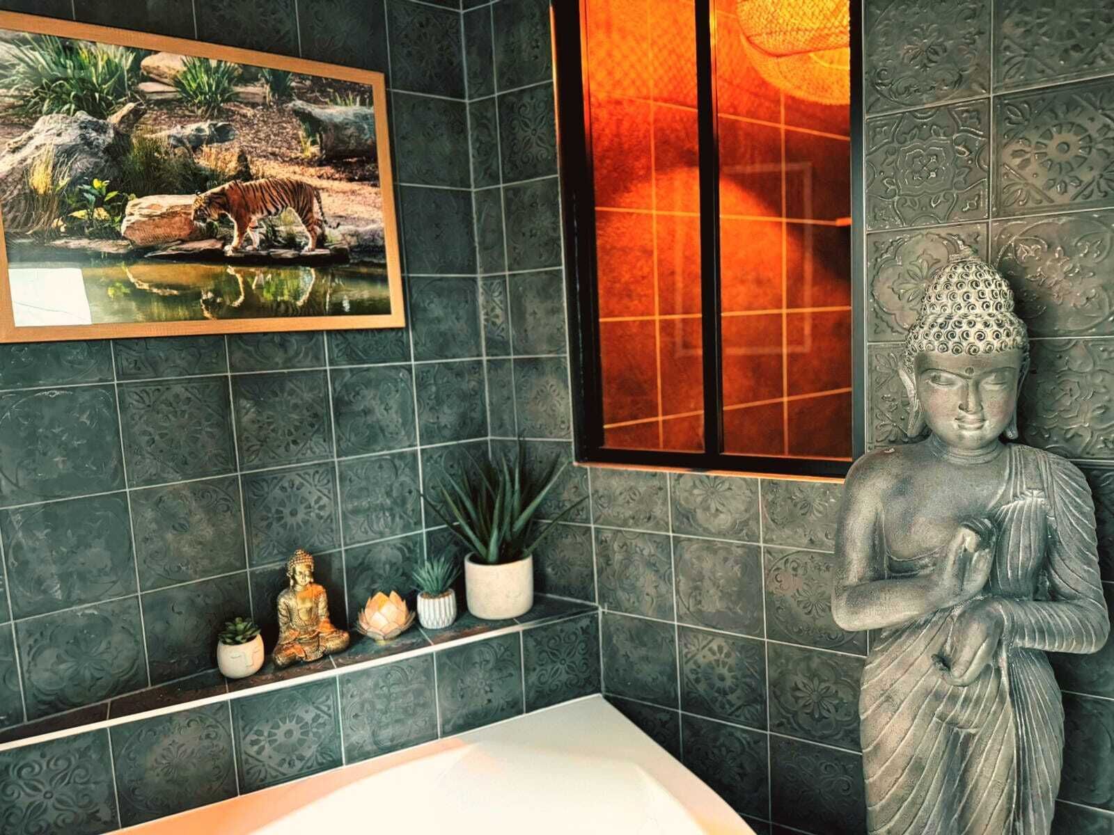 Een badkamer met donkere, patroonrijke tegels, een Boeddha-beeldje, een ingelijst beeld van een tijger en kleine decoratieve items op een plank.