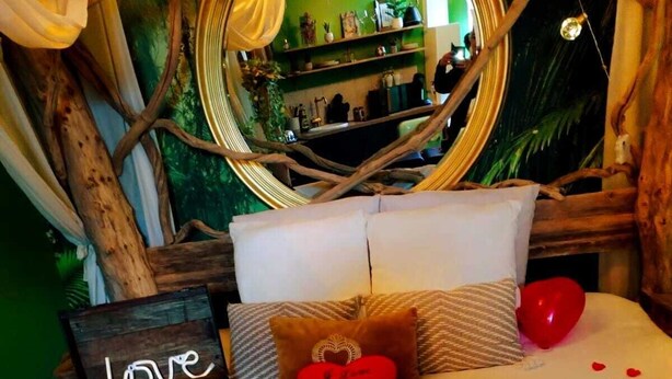 Una cama con un arreglo de pétalos de rosa en forma de corazón, una botella de champán y una pequeña cesta con una botella de vino Raffaello.