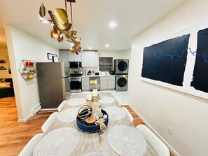Una cocina moderna con electrodomésticos de acero inoxidable, una zona de comedor con una mesa redonda y sillas blancas, y un ventanal con un cuadro abstracto en tonos negros.