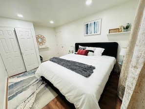 Un dormitorio con una cama, paredes blancas, una puerta, un espejo y un cuadro.