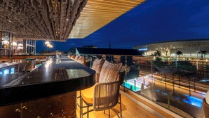 A bar area with a pool view, modern lighting, and a large building in the background. - Zaria Court Kigali, Tapestry Collection by Hilton (Kigali)