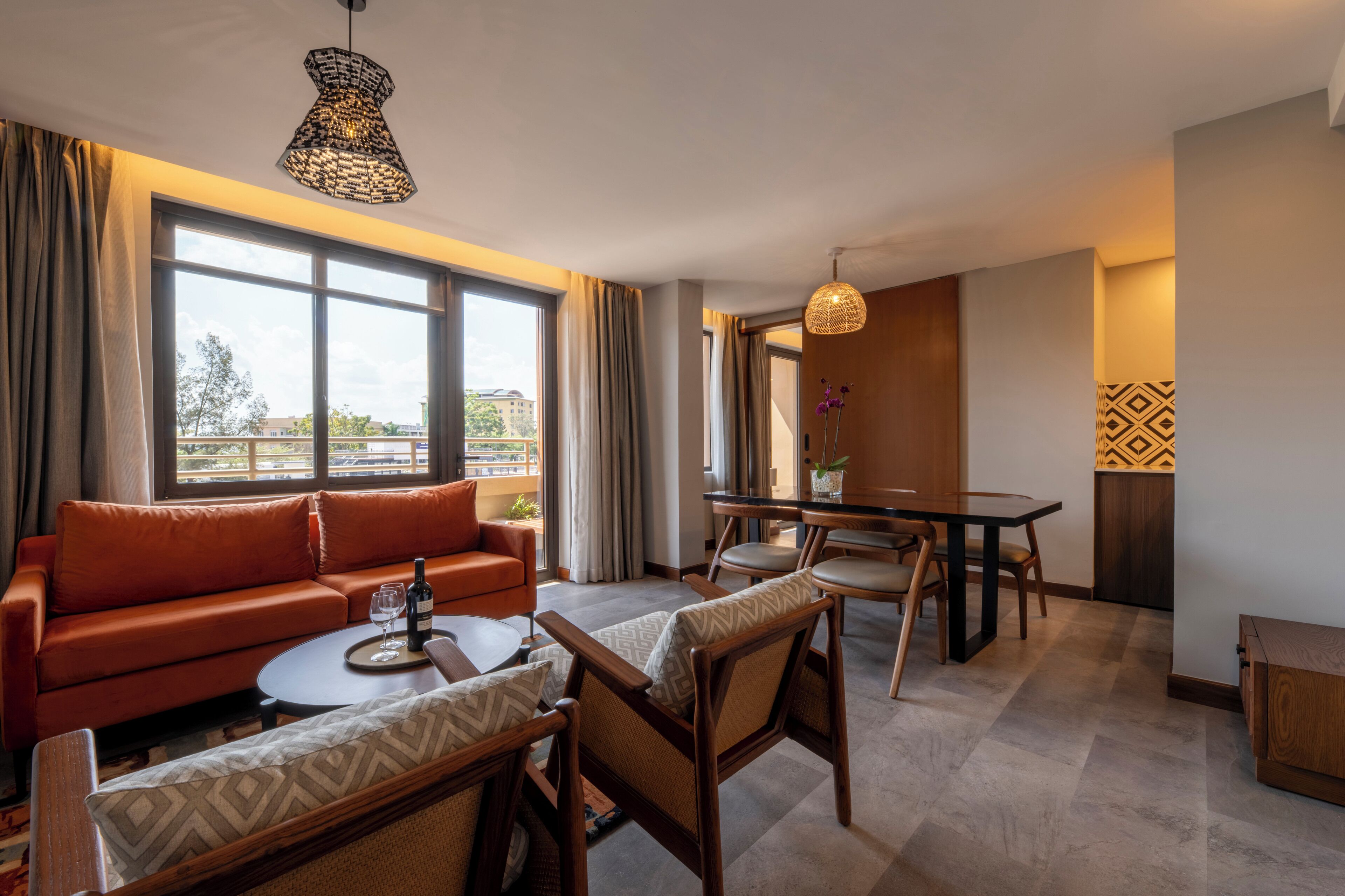 A modern hotel room with a sofa, chairs, a dining table, and a large window offering a view.