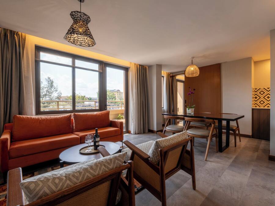 A modern hotel room with a sofa, chairs, a dining table, and a large window offering a view.