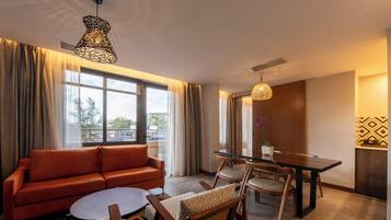 A modern hotel room with a sofa, dining table, chairs, a round coffee table, and large windows with curtains.
