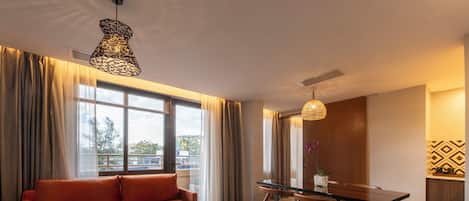 A modern hotel room with a sofa, dining table, chairs, a round coffee table, and large windows with curtains.