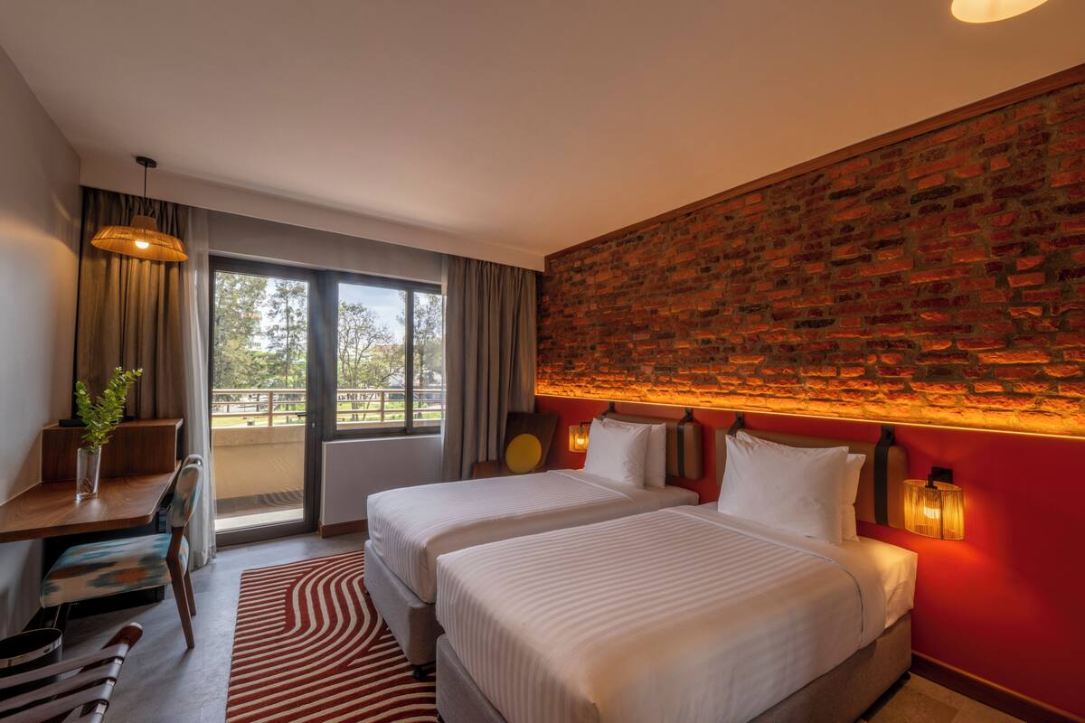 A hotel room with two beds, a desk, a chair, a window with curtains, and a brick wall.