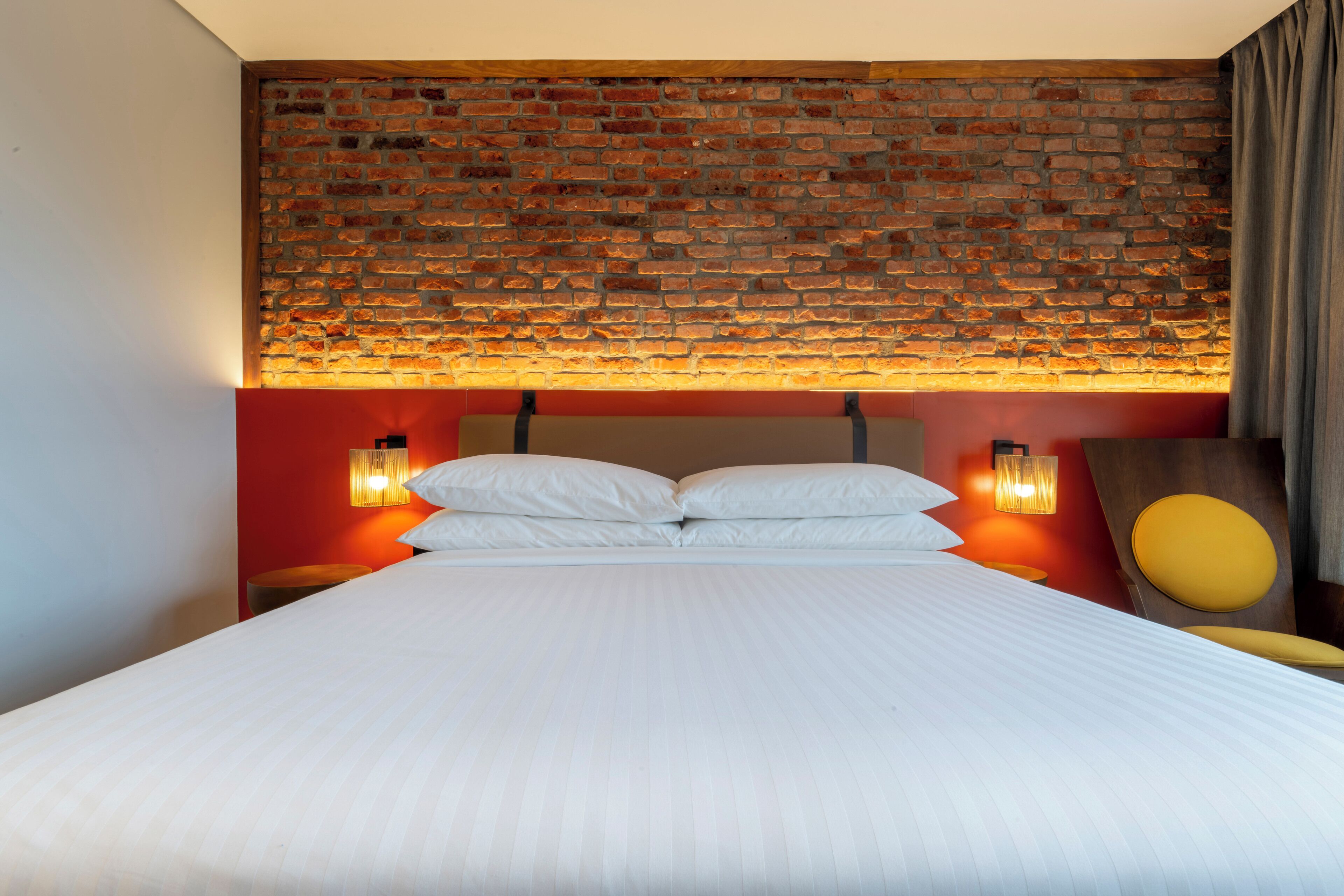 Una habitación de hotel con pared de ladrillo, una cama con ropa blanca, dos lámparas de noche y una silla amarilla.