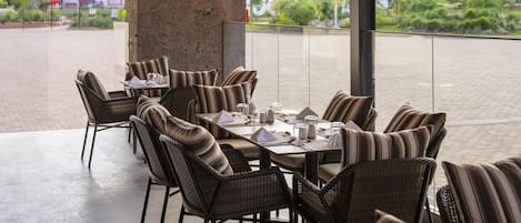 An outdoor dining area with wicker chairs and tables, a hanging light fixture, and a view of buildings and greenery.