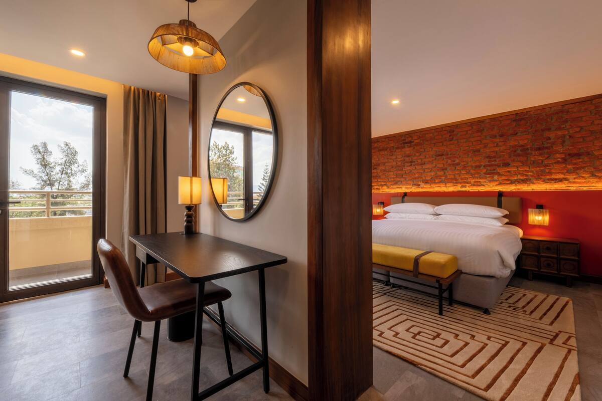 A hotel room with a bed, a desk with a chair, a large mirror, a window with curtains, and a brick wall.
