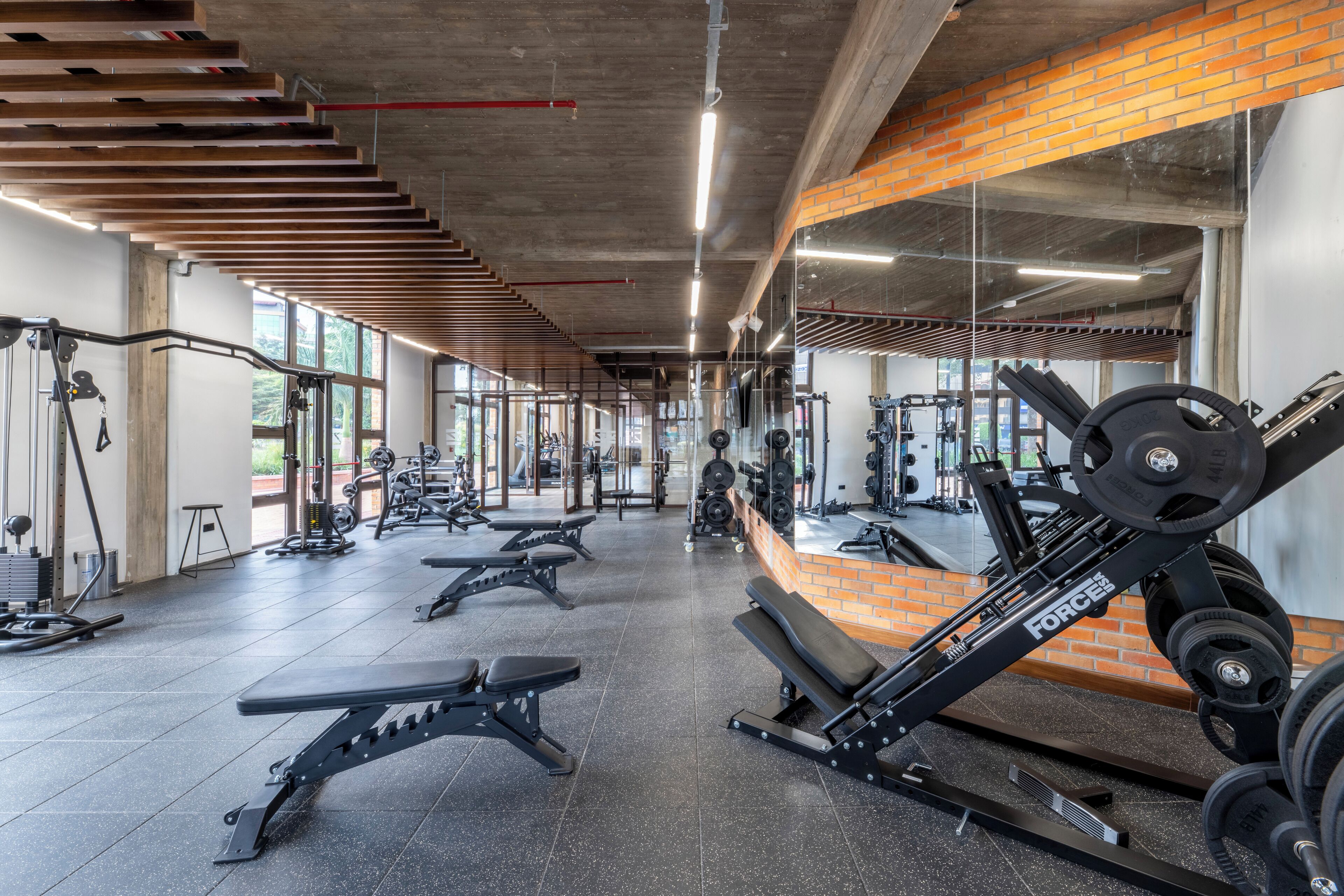 Un gimnasio con diverso equipo de ejercicio, ventanas amplias y una pared de ladrillo.
