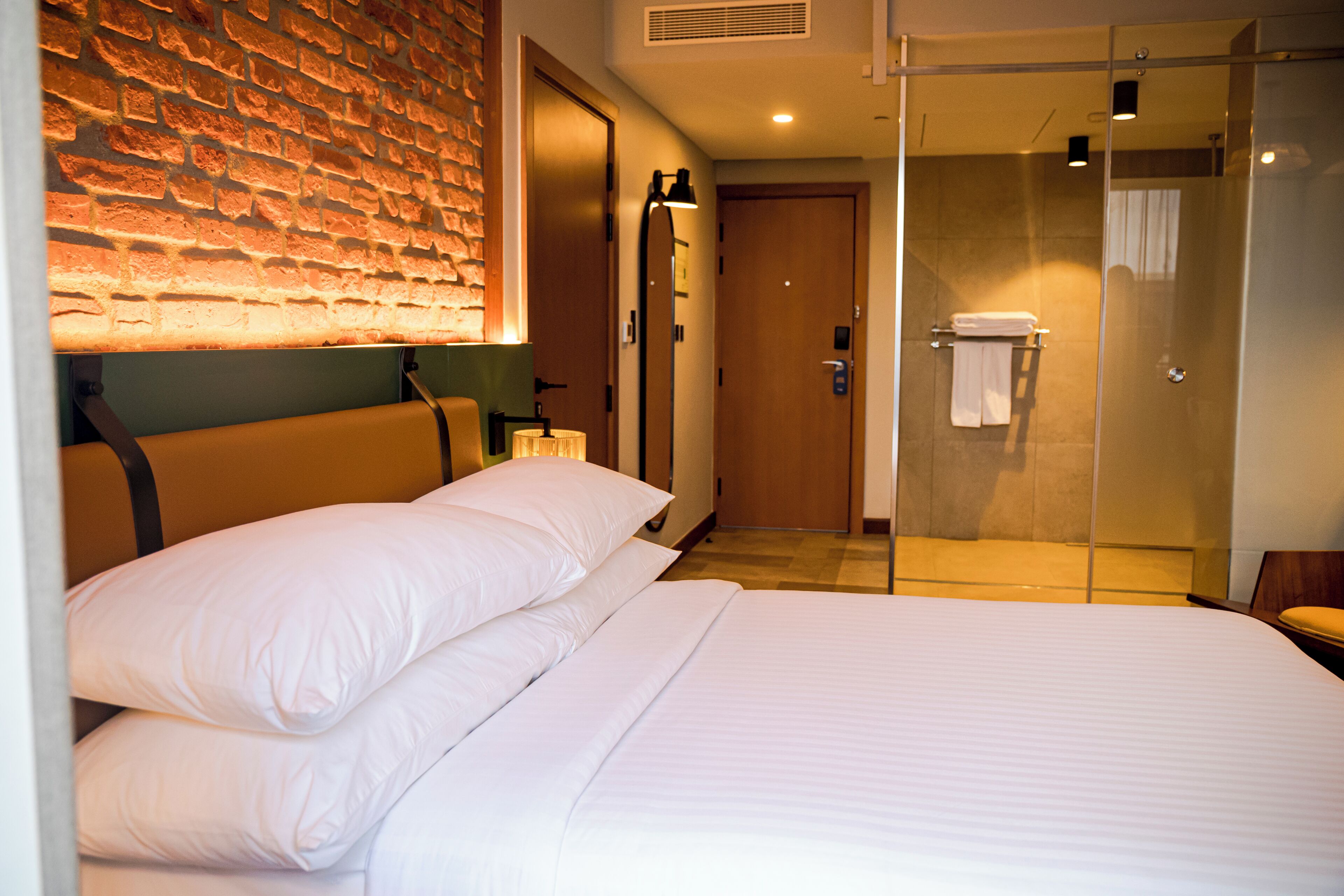 A hotel room with a brick wall, a bed with white linens, a glass shower enclosure, and a wooden door.