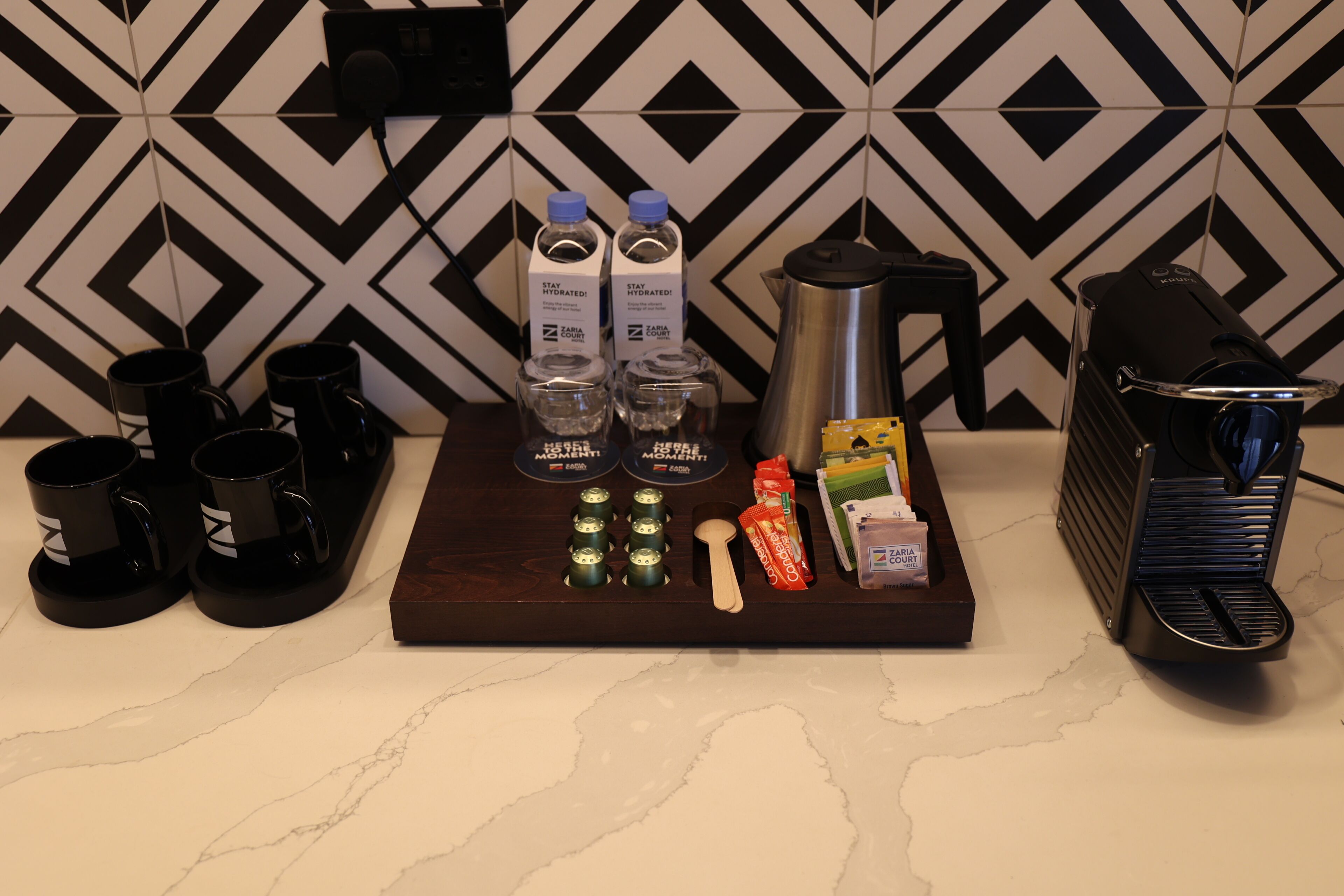 A kitchen counter with a coffee maker, two water bottles, a coffee pot, a spoon, a box of tissues, and a small trash bin.