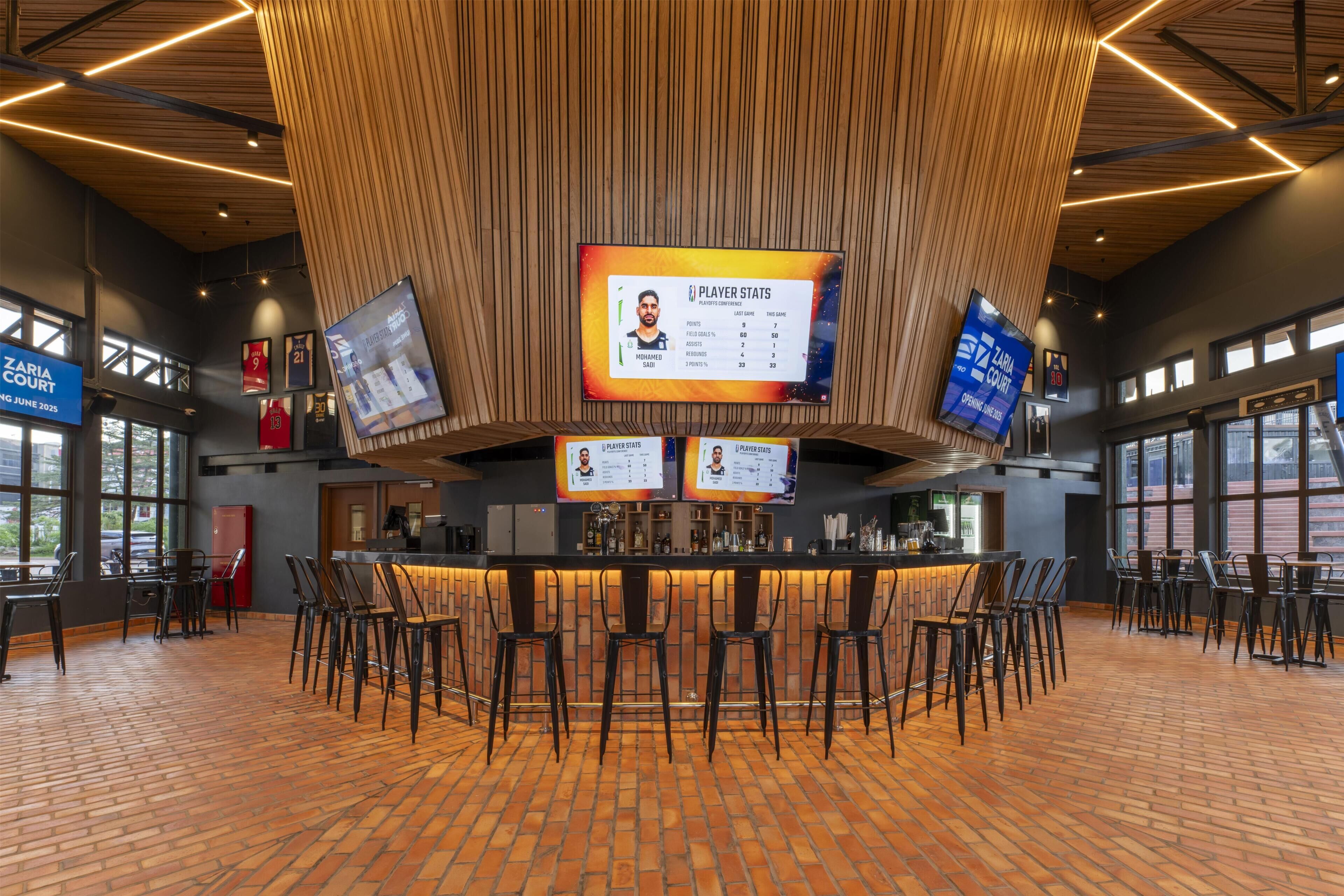A sports bar with a central counter, multiple screens displaying player stats, and a brick floor.